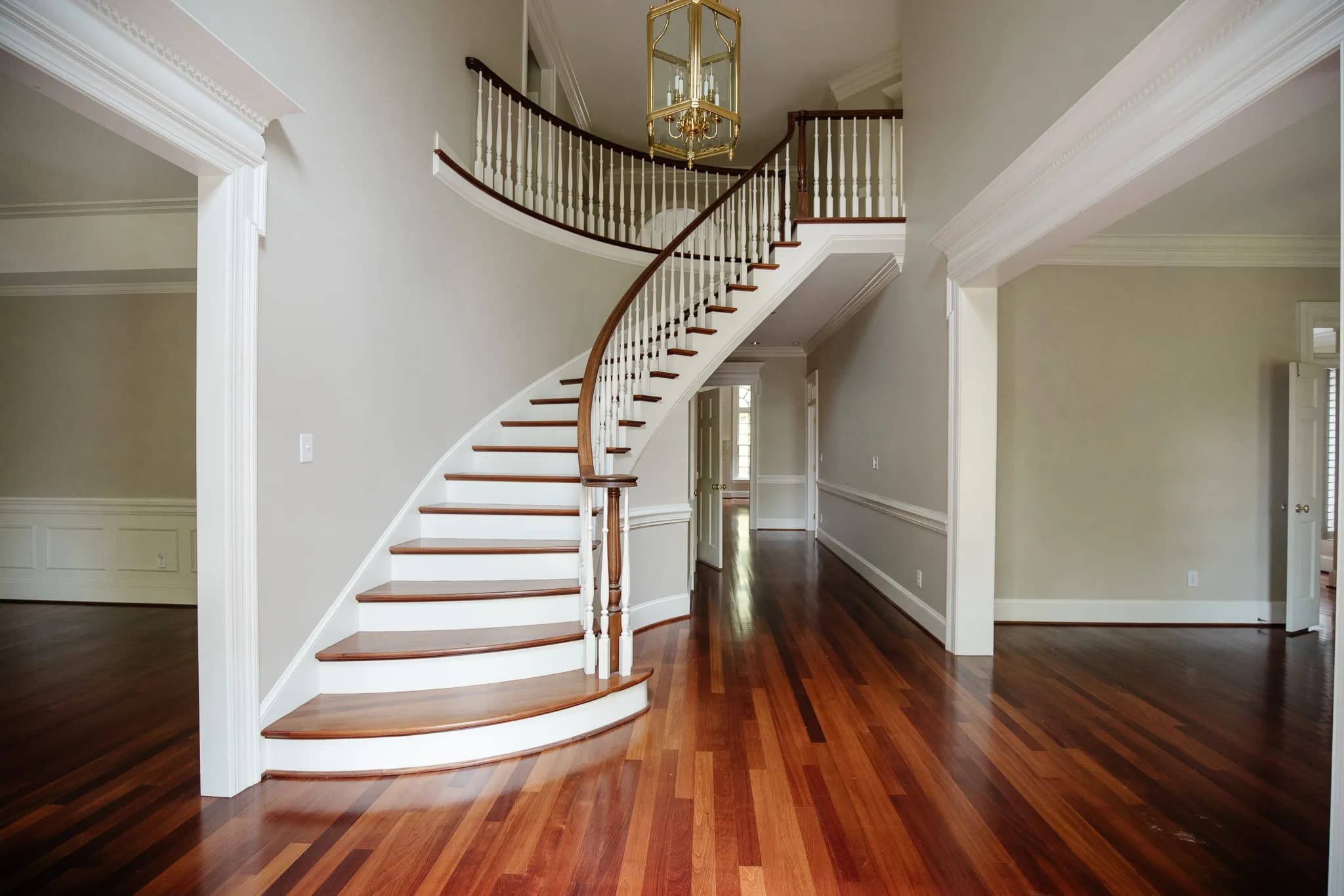 Curved staircase with white painted balusters, dark wood handrail and cherry hardwood floors, lit by a brass lantern chandelier